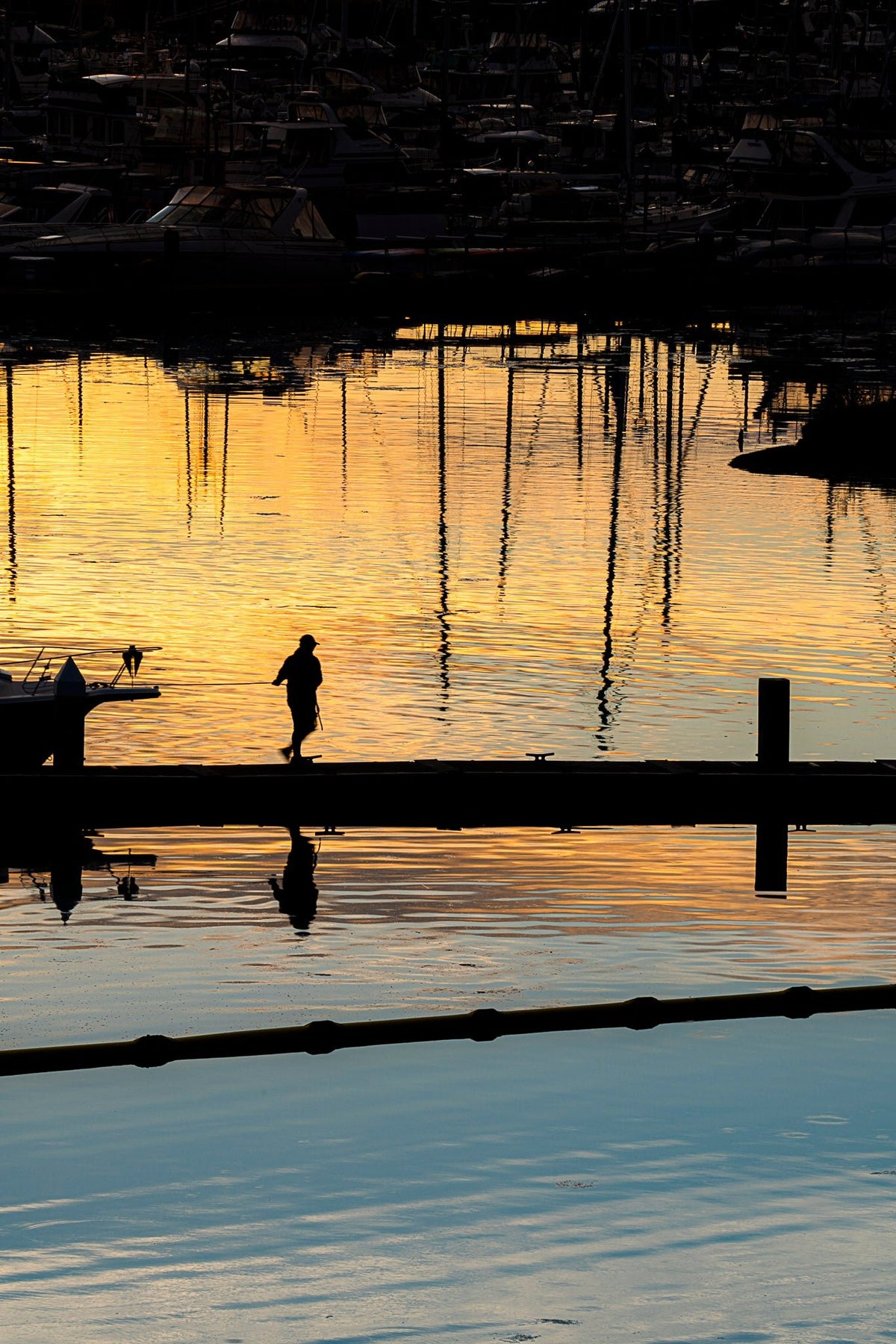 00_30 by Nick Patterson. A silhouetted figure and sailboat masts reflected in golden water.